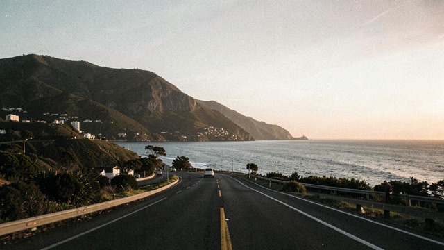A curving coastal road between a hillside and the ocean in soft light.