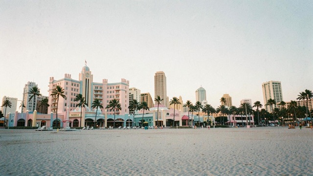 An open sandy beach in front of pastel buildings and palm trees.