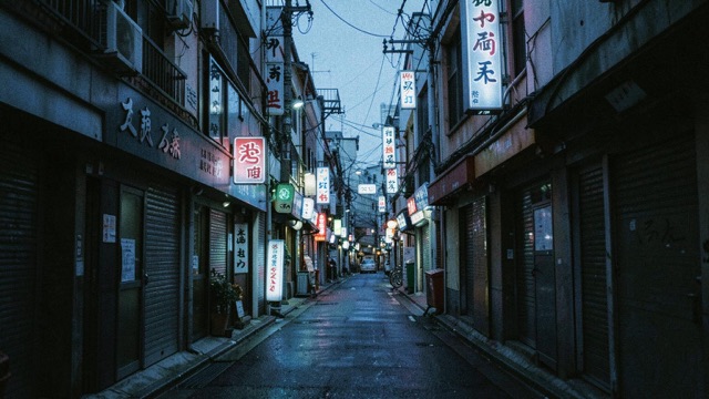 A narrow urban street with hanging signs and wet paving in cool light.