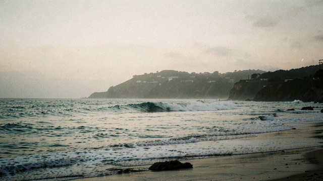 Small waves approaching a sandy beach with a headland in the distance.