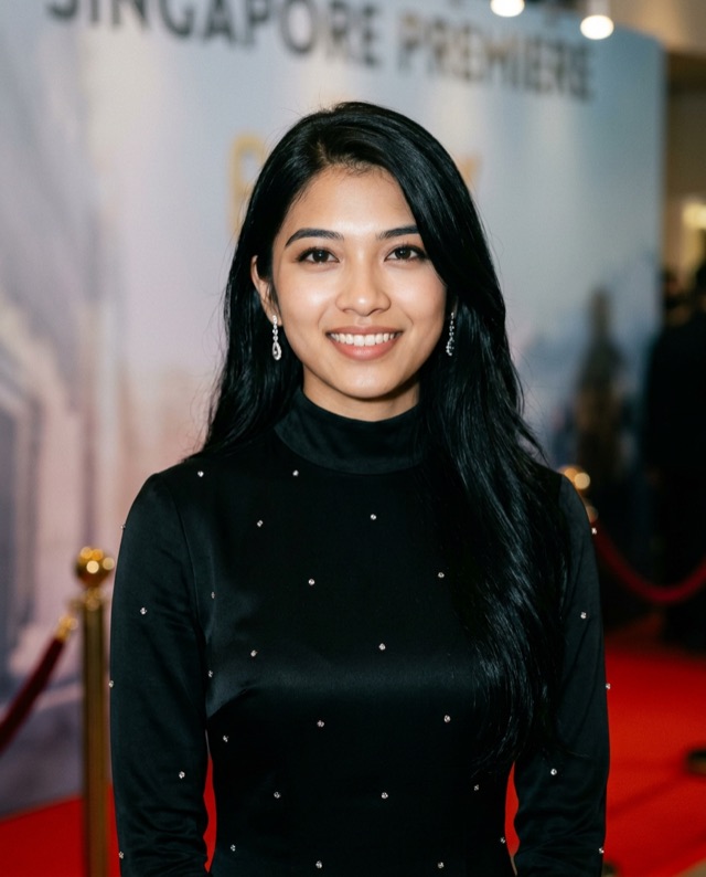 Priya smiling in a black outfit on a red carpet in front of an event backdrop.